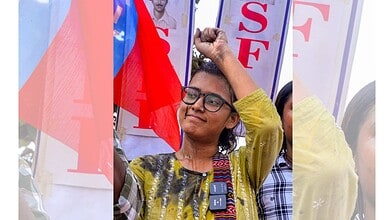 Young woman with glasses raising her fist at a protest or rally with banners in the background.