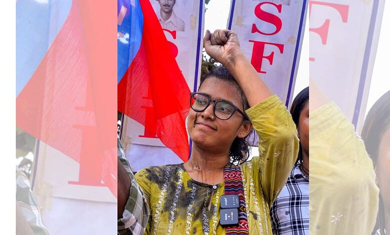 Young woman with glasses raising her fist at a protest or rally with banners in the background.