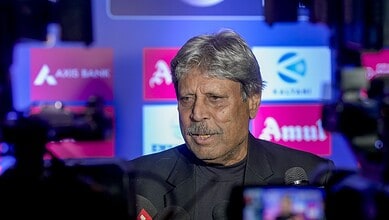 Kapil Dev speaking at a press event with a backdrop of sponsor logos.