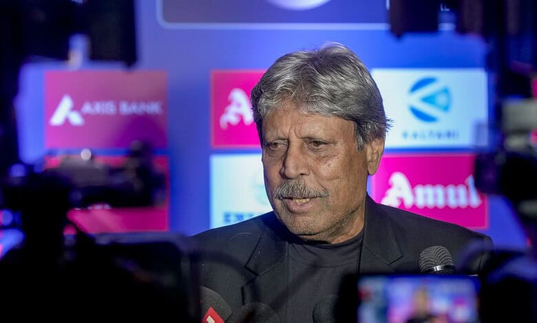 Kapil Dev speaking at a press event with a backdrop of sponsor logos.