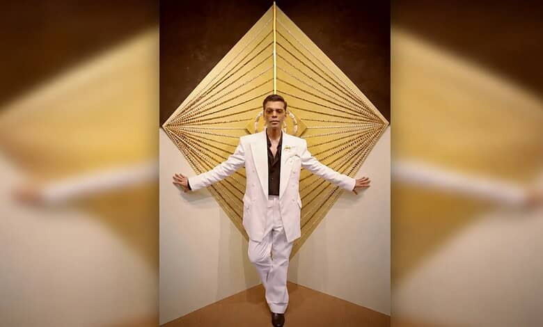 Celebrity posing in white suit against geometric background for Koffee With Karan season 9.