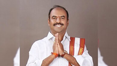 Smiling man in white shirt with traditional Indian shawl, hands folded in greeting.