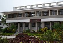 Modern library building with greenery and flowers in front, under a cloudy sky.
