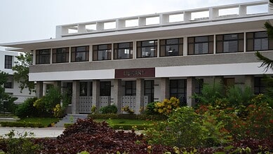 Modern library building with greenery and flowers in front, under a cloudy sky.