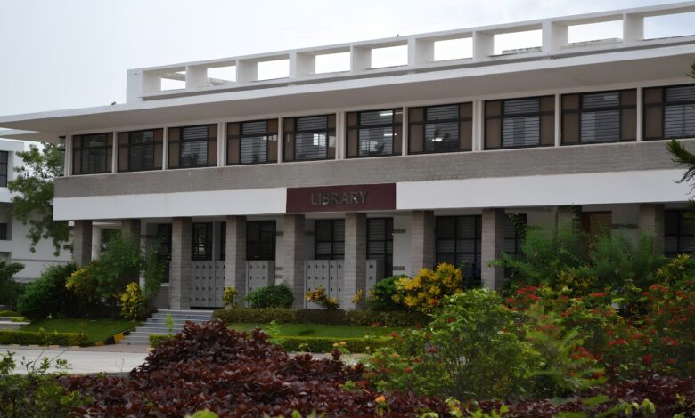 Modern library building with greenery and flowers in front, under a cloudy sky.