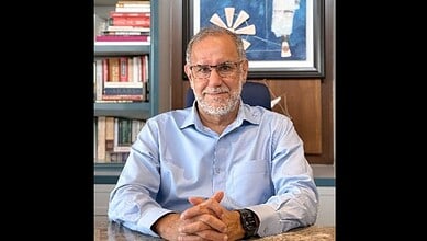 Ex-diplomat in office, wearing glasses and a light blue shirt, with books and artwork in the background.