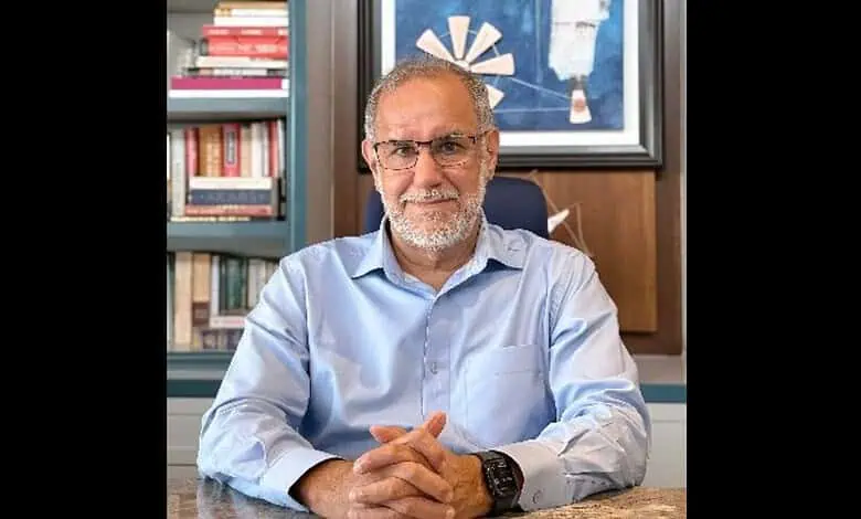 Ex-diplomat in office, wearing glasses and a light blue shirt, with books and artwork in the background.
