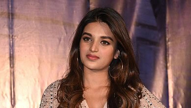 Nidhi Agarwal at a public event, wearing a beige outfit with embellishments and hoop earrings.