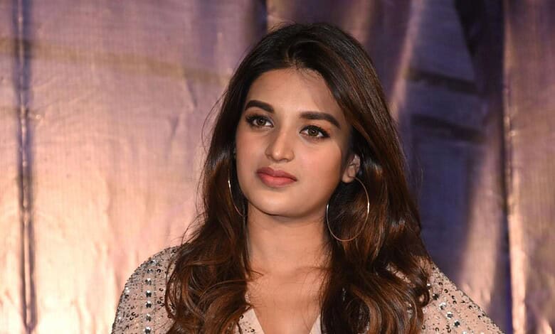 Nidhi Agarwal at a public event, wearing a beige outfit with embellishments and hoop earrings.