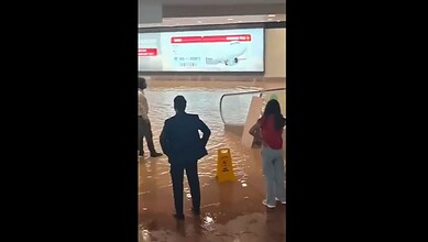 People walking through waterlogged area at Noida airport after heavy rain, with a warning sign visible.