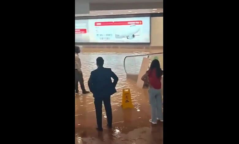 People walking through waterlogged area at Noida airport after heavy rain, with a warning sign visible.