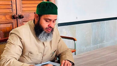 Man with a beard wearing a green cap and beige traditional attire sitting at a wooden table.