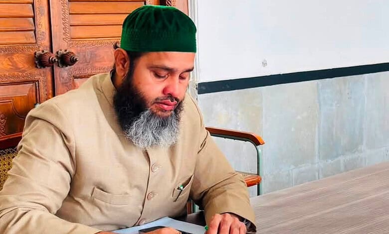 Man with a beard wearing a green cap and beige traditional attire sitting at a wooden table.