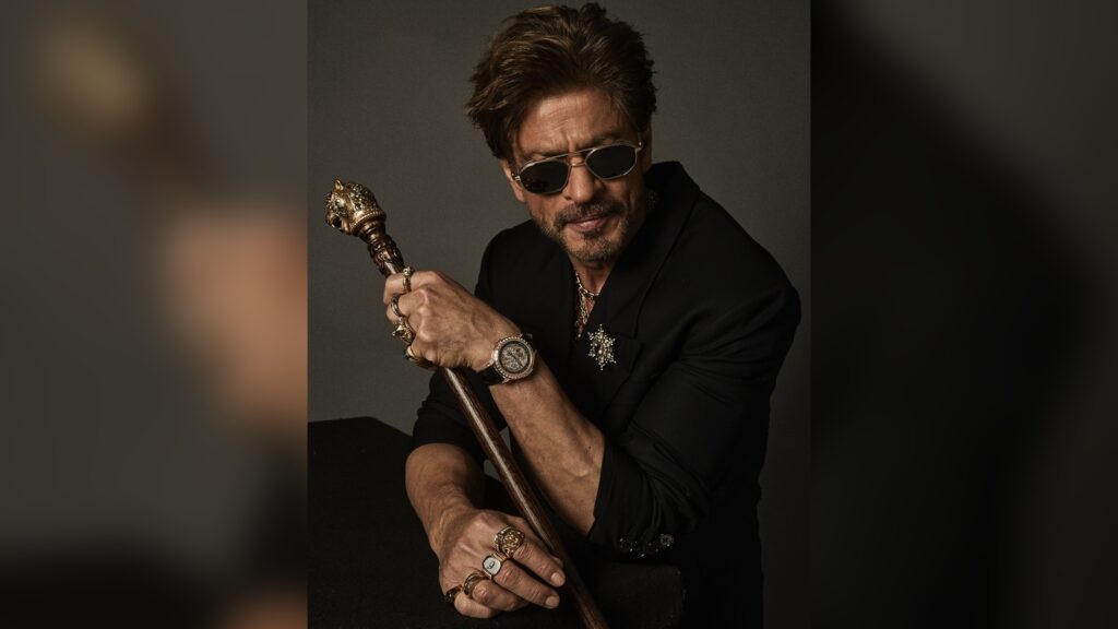 Shah Rukh Khan in a stylish black outfit with sunglasses, holding a cane, posing confidently.