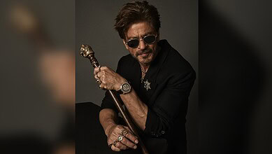 Shah Rukh Khan in a stylish black outfit with sunglasses, holding a cane, posing confidently.