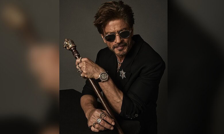 Shah Rukh Khan in a stylish black outfit with sunglasses, holding a cane, posing confidently.