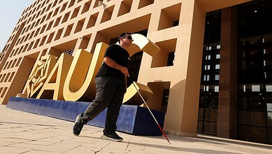 Blind Egyptian man with white cane walking past a modern building with large yellow letters.