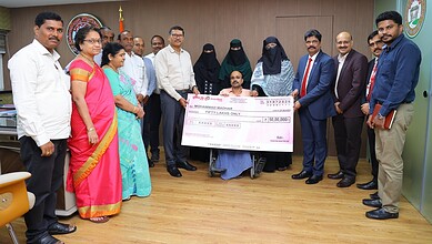 Group of officials and family members present a cheque to a man with a leg injury in an office setting.