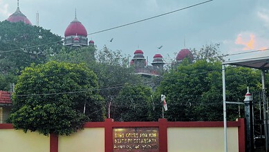 Historical building with red domes and lush greenery in front, under a cloudy sky.