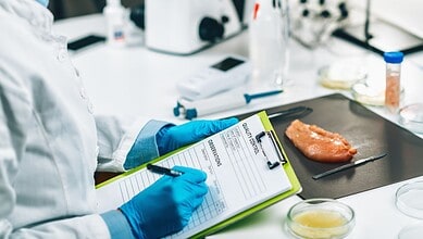Food testing laboratory with a scientist recording results and chicken sample on a black tray.