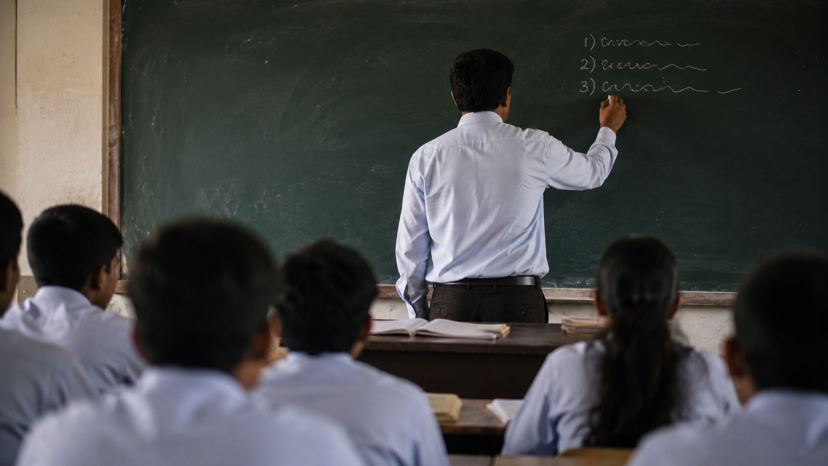 Teacher writing on blackboard in a classroom with students in white uniforms.