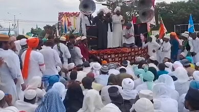 Large crowd gathered at a public event with speakers and stage, many wearing traditional attire and turb.