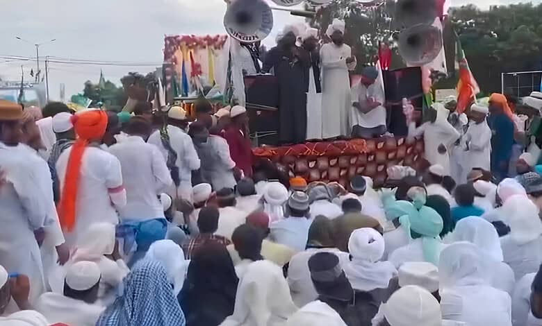 Large crowd gathered at a public event with speakers and stage, many wearing traditional attire and turb.