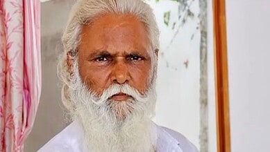 Elderly man with white hair and beard wearing white traditional attire, serious expression.