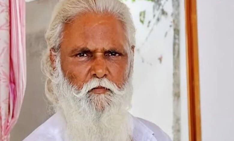 Elderly man with white hair and beard wearing white traditional attire, serious expression.