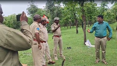 Man in blue shirt speaking to police officers in a park with trees and grass.