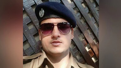 Young police officer wearing sunglasses and a beret in front of a wooden fence.