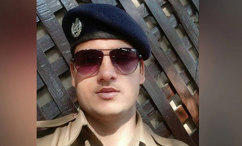 Young police officer wearing sunglasses and a beret in front of a wooden fence.