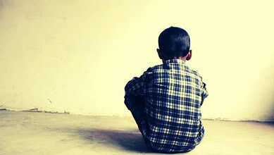 Child sitting alone against a plain wall, facing away from the camera.