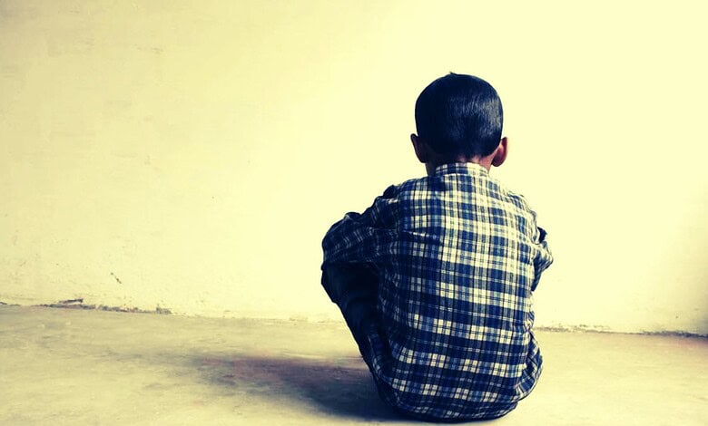 Child sitting alone against a plain wall, facing away from the camera.