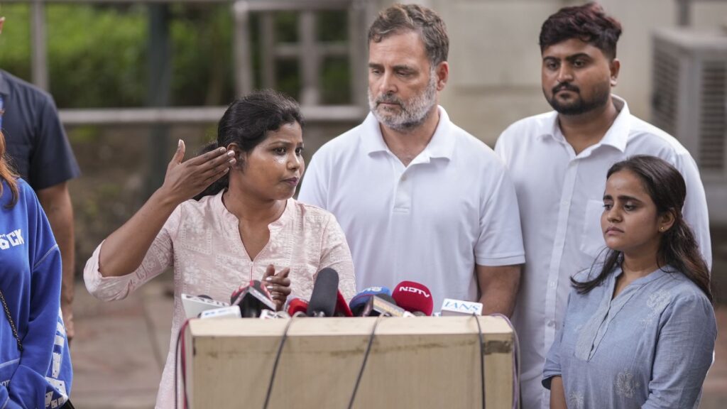 Indian woman speaking at a press conference with microphones, surrounded by men and women.