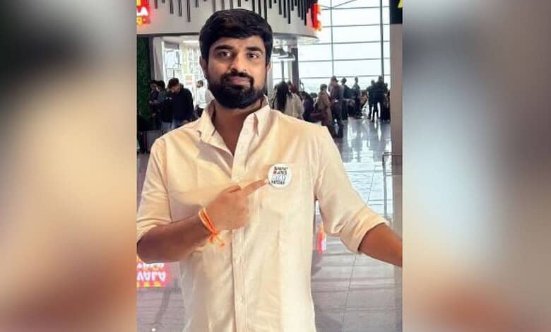 Naveen Pettem at an airport, wearing a white shirt and pointing to a badge.