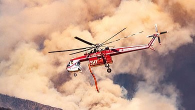 Red firefighting helicopter flying through smoky sky during wildfire response.