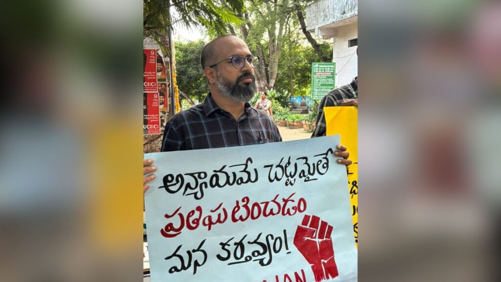 Man holding a protest sign with Telugu text and a red fist symbol outdoors.