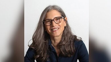 Smiling woman with long grey hair and glasses in a dark blazer against a plain background.