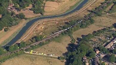 Aerial view of a derailed train near a river in England, with emergency responders present.
