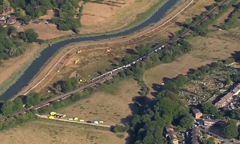 Aerial view of a derailed train near a river in England, with emergency responders present.