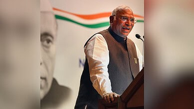 Kharge speaking at a podium during a political event, with a large banner in the background.