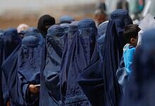 Afghan girls in blue burqas gather outdoors, some with children, under a clear sky.