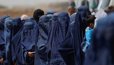 Afghan girls in blue burqas gather outdoors, some with children, under a clear sky.
