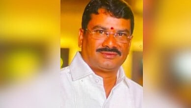 Man in white shirt with glasses and red tilak, smiling at camera.