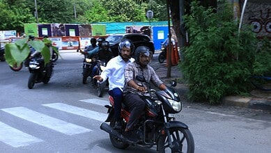 Two men riding a black scooter at KBR Park traffic in Hyderabad.