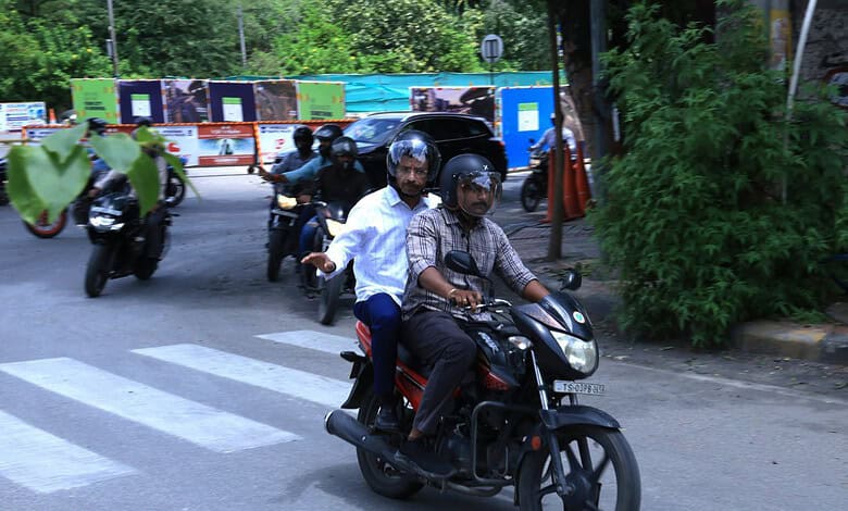 Two men riding a black scooter at KBR Park traffic in Hyderabad.
