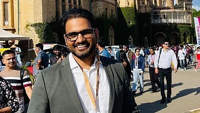 Smiling man in sunglasses and suit at outdoor event with historic building background.