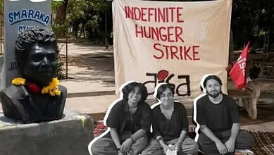 Protesters with a banner for indefinite hunger strike and a bust statue in the background.