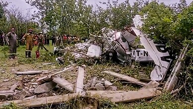 Wreckage of a crashed aircraft in a rural area with rescue workers and onlookers present.